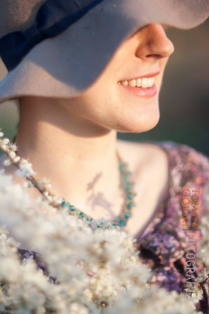 Portret kobiety w kapeluszu, z uśmiechem i turkusowym naszyjnikiem, otoczonej kwitnącymi gałązkami drzewa.