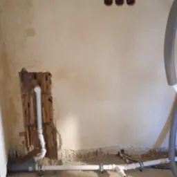 Surowe wnętrze w trakcie remontu z odsłoniętymi rurami instalacji wodnej i grzewczej w ścianach, widoczne przewody w izolacji termicznej i fragmenty instalacji podłogowej.