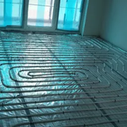 Białe rurki układane w pętle na siatce podłogowej, instalacja ogrzewania podłogowego w surowym pomieszczeniu z oknami zabezpieczonymi folią.