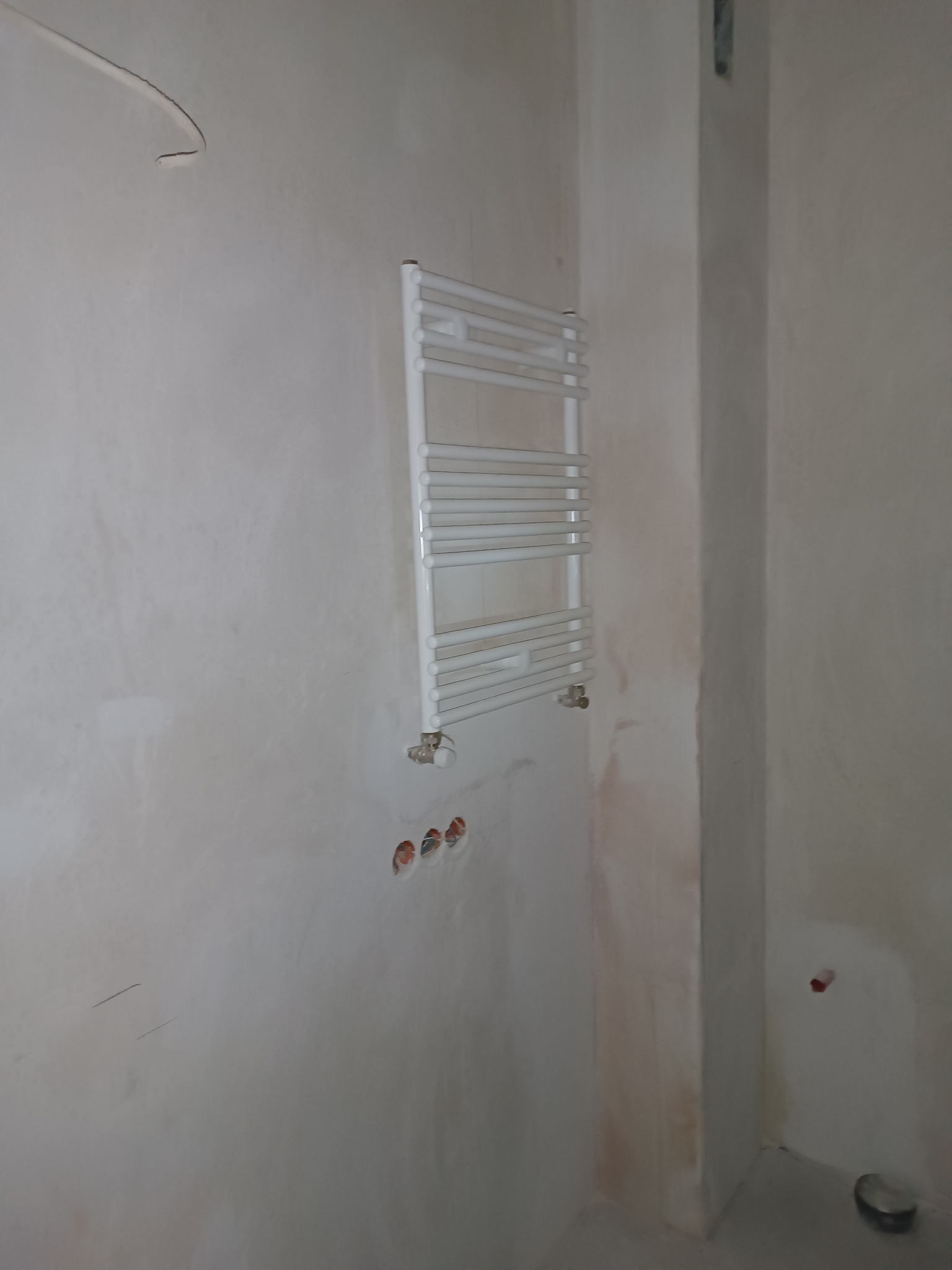 Biały grzejnik łazienkowy drabinkowy zamontowany na surowej ścianie w trakcie remontu, widoczne otwory pod instalację hydrauliczną.