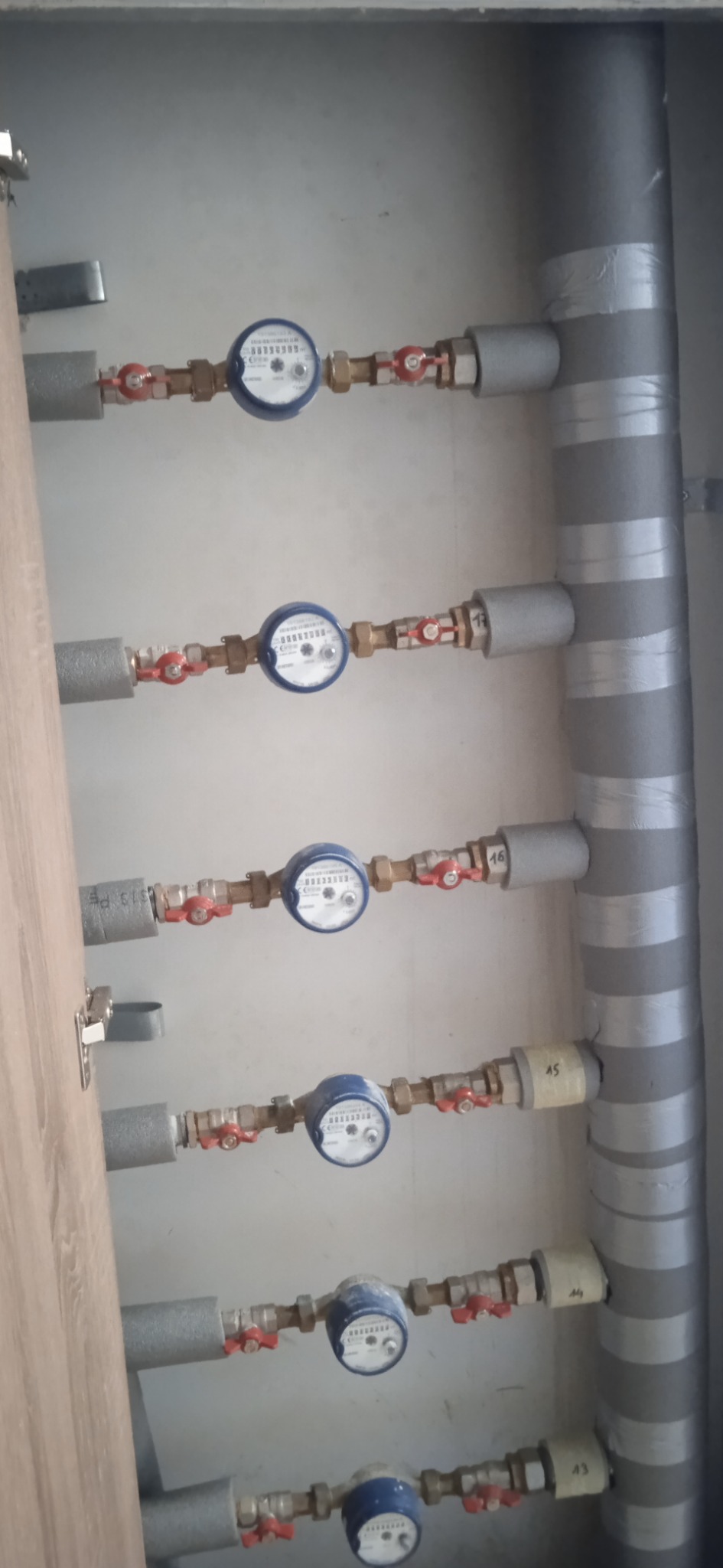 Rzędy liczników wody z zaworami i izolacją termiczną na rurach, zamontowane w szafce z drewnianymi drzwiczkami.