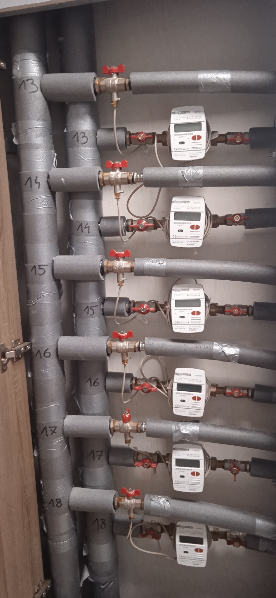 Rozdzielacz instalacji grzewczej z izolowanymi rurami oznaczonymi numerami od 13 do 18, zaworami z czerwonymi pokrętłami i licznikami ciepła, widoczne w szafce.