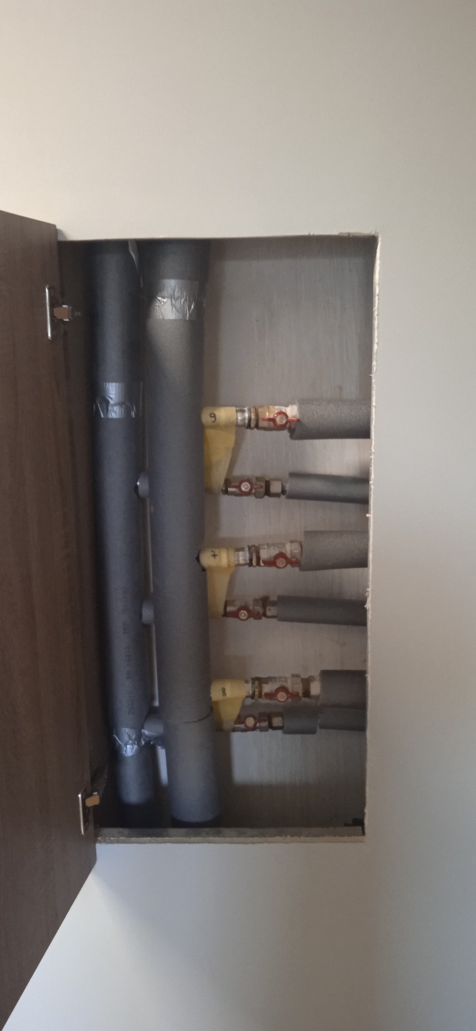 Ukryte w szafce pionowe rury grzewcze z izolacją i zaworami odcinającymi, zabezpieczone taśmą, instalacja po montażu.