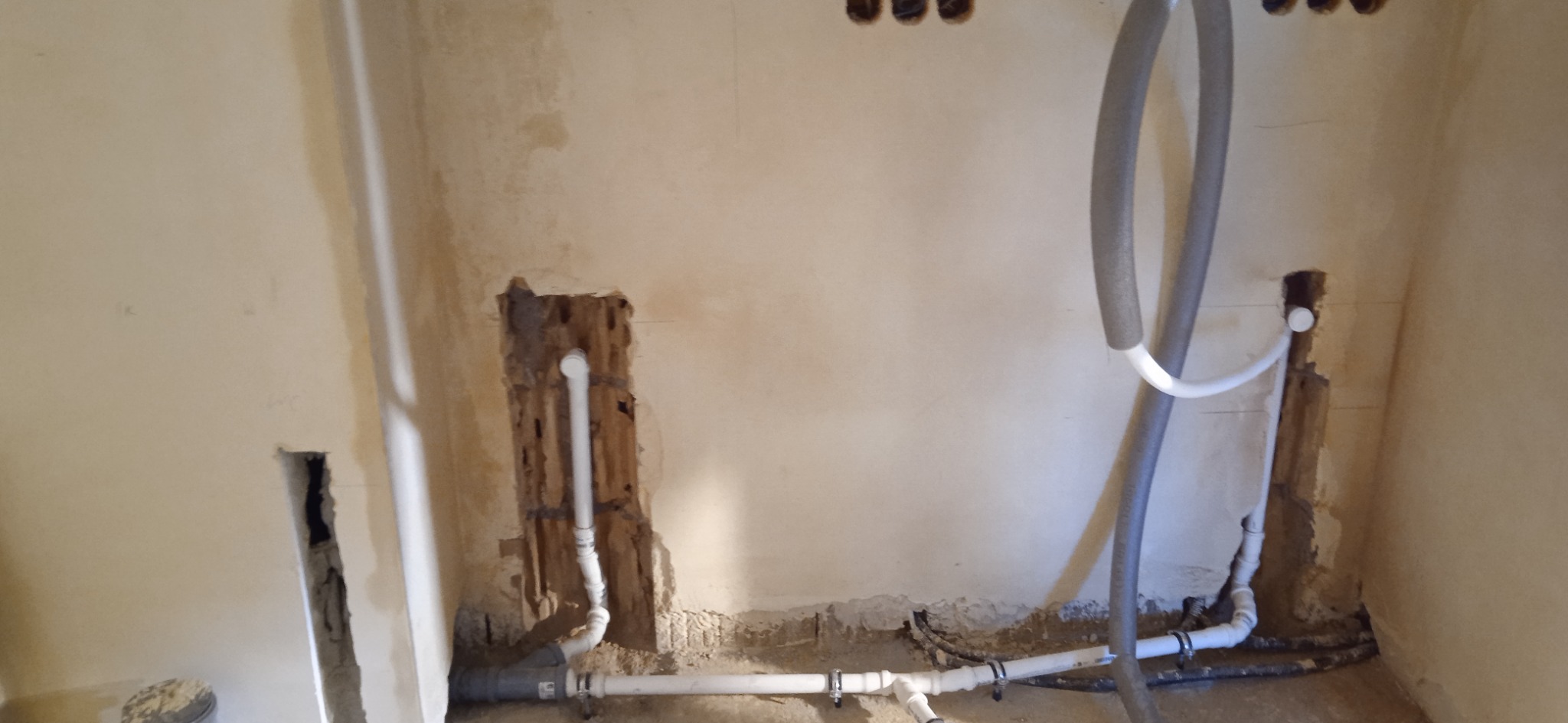 Surowe wnętrze w trakcie remontu z odsłoniętymi rurami instalacji wodnej i grzewczej w ścianach, widoczne przewody w izolacji termicznej i fragmenty instalacji podłogowej.