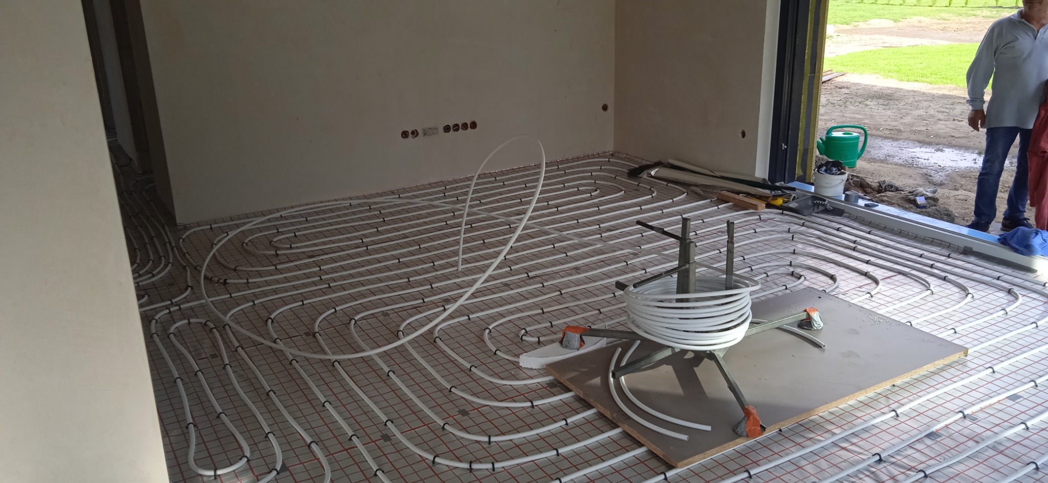 Białe rury układane w pętle na czerwonej siatce, przygotowanie do wylewki podłogowej, widoczny rozwijak do rur i fragment osoby w tle.
