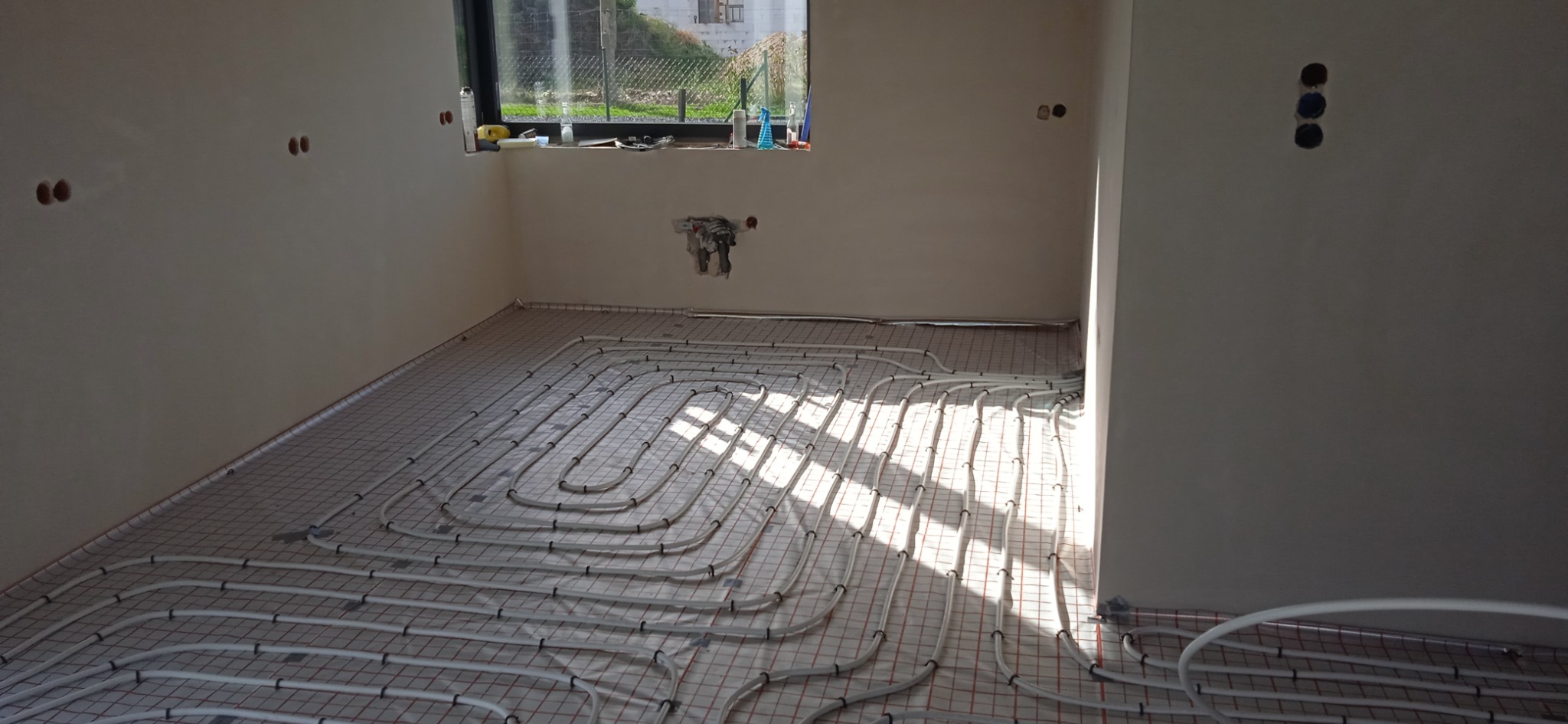 Białe rury systemu ogrzewania podłogowego ułożone na siatce w surowym pomieszczeniu z oknem, widoczne otwory na gniazdka elektryczne i instalację hydrauliczną.