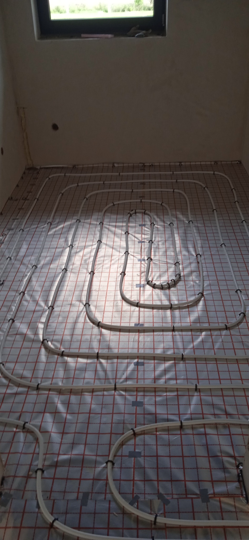 Białe rury ułożone w pętle na folii z czerwoną kratką, przygotowane do zalania posadzki w pomieszczeniu z oknem u góry.