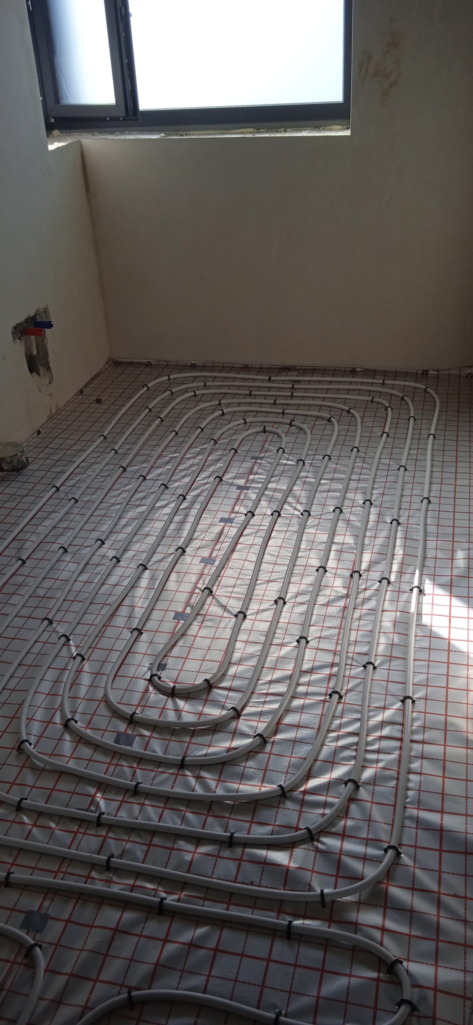 Białe rurki systemu ogrzewania podłogowego ułożone w pętle na folii z czerwoną siatką, widok z góry w surowym wnętrzu z oknem dachowym.
