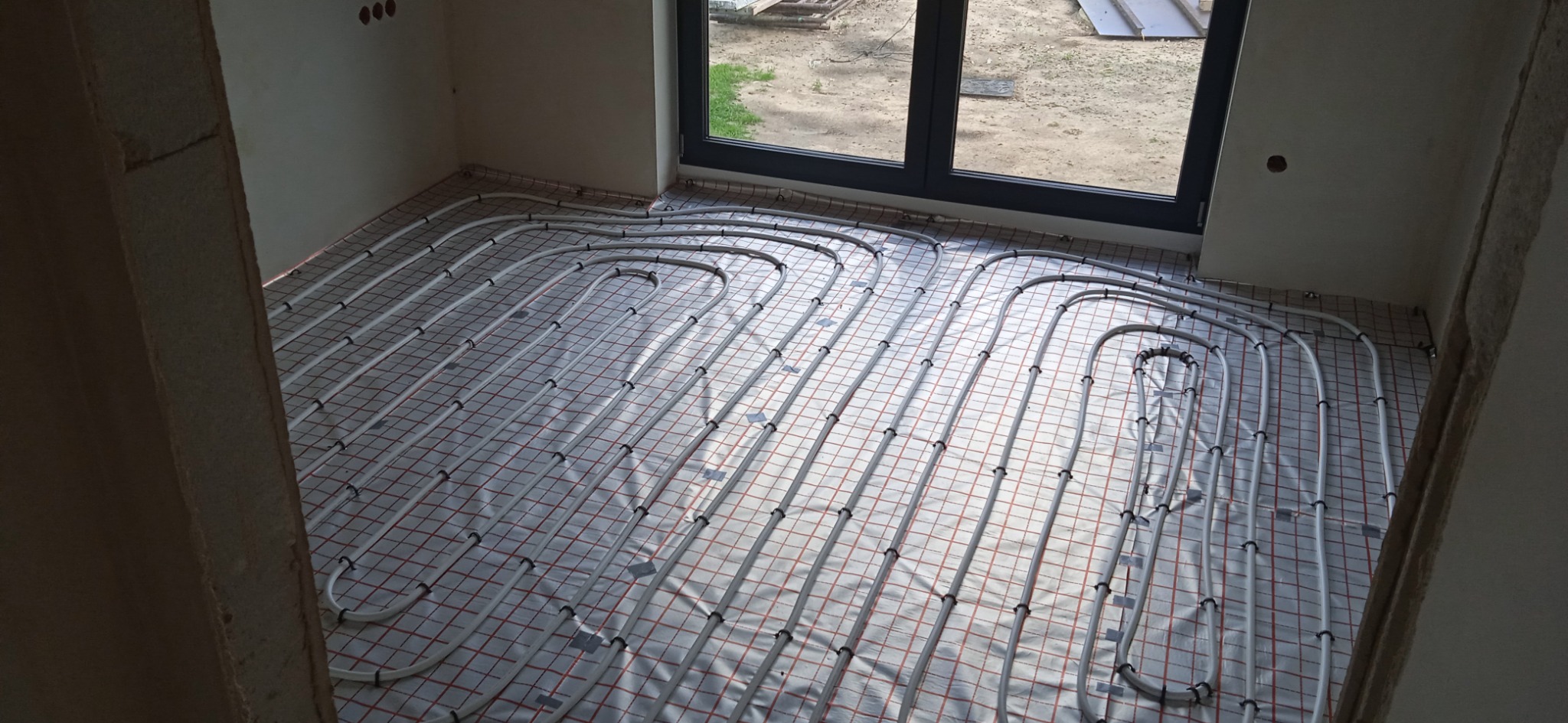 Rozłożone rury systemu grzewczego na folii z siatką w surowym wnętrzu przed wylaniem posadzki, widoczne okno z widokiem na zewnątrz.