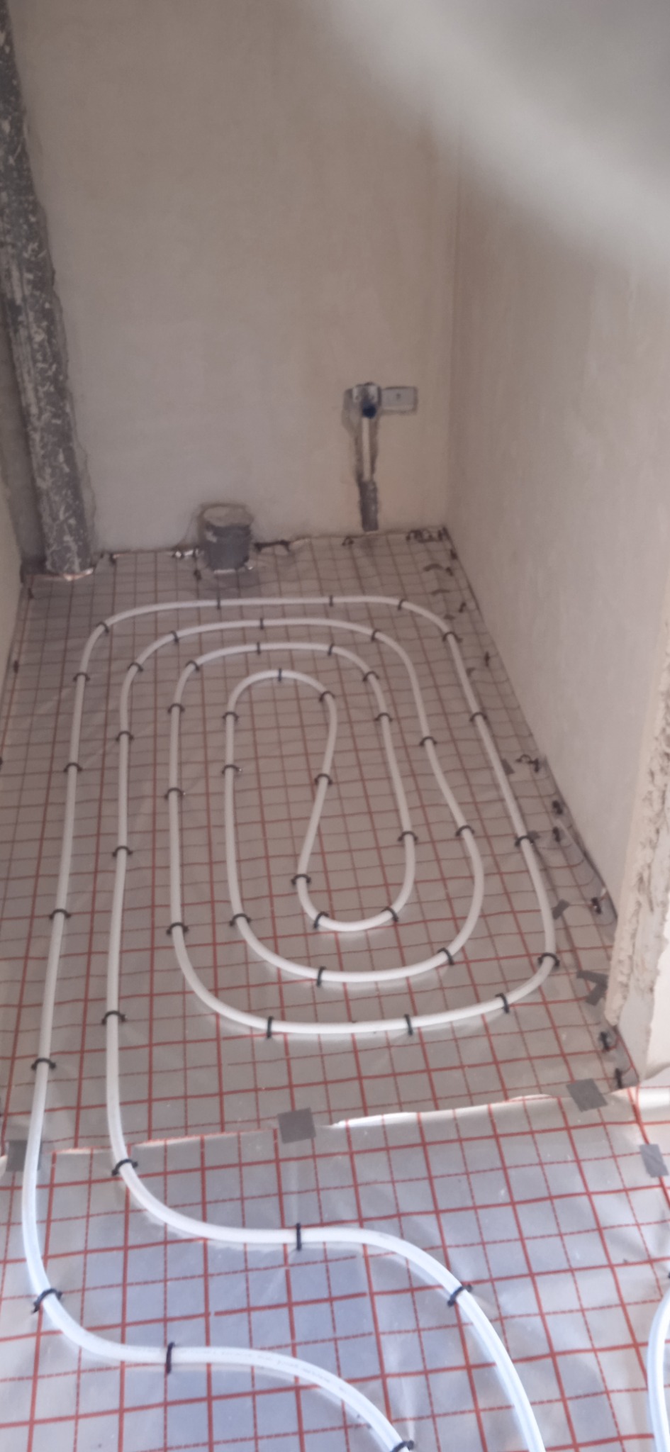 Białe rury ogrzewania podłogowego ułożone na folii z czerwoną kratką w surowym pomieszczeniu podczas montażu, widoczne mocowania rur do folii.
