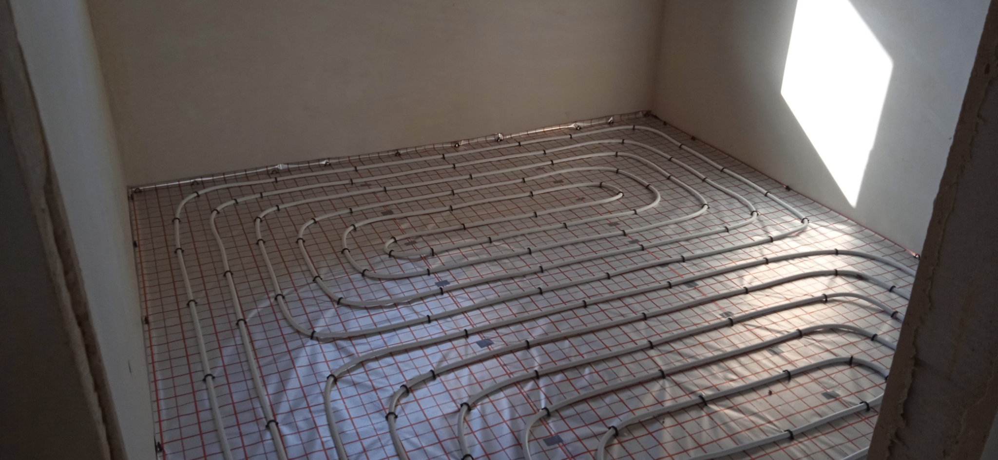 Białe rury systemu ogrzewania podłogowego ułożone na folii z czerwoną siatką w surowym, jasnym pomieszczeniu, widoczne czarne uchwyty mocujące rury.