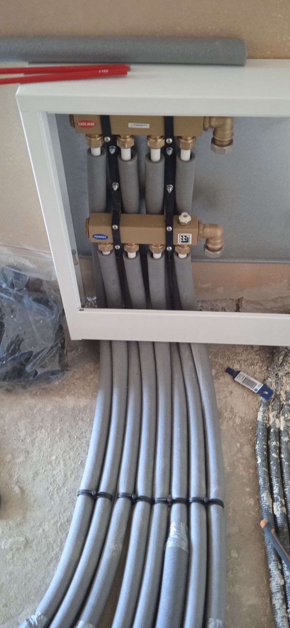 Rozdzielacz instalacji ogrzewania podłogowego w białej szafce, rury w szarej izolacji termicznej, widoczne elementy mosiężne i czarne opaski zaciskowe, surowy beton na ścianie w tle.