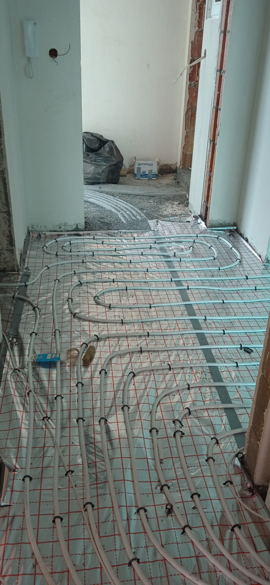 Białe rurki systemu ogrzewania podłogowego ułożone na srebrnej folii z czerwoną kratką, zabezpieczone czarnymi klipsami, widok na surową podłogę w tle, worek z gruzem i karton po materiałach...