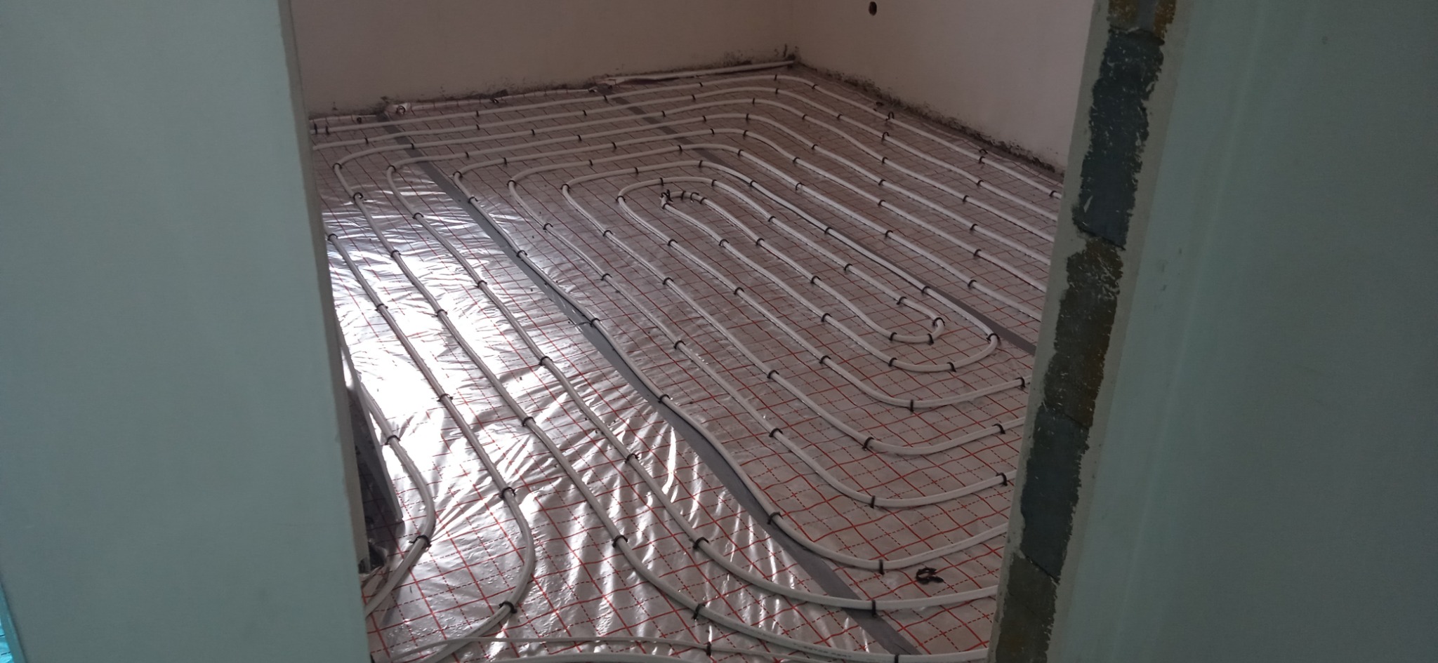 Białe rurki systemu rozprowadzenia ciepła ułożone na srebrnej folii z czerwoną siatką, widok z góry na podłogę w surowym stanie, w tle ściany w jasnym odcieniu.
