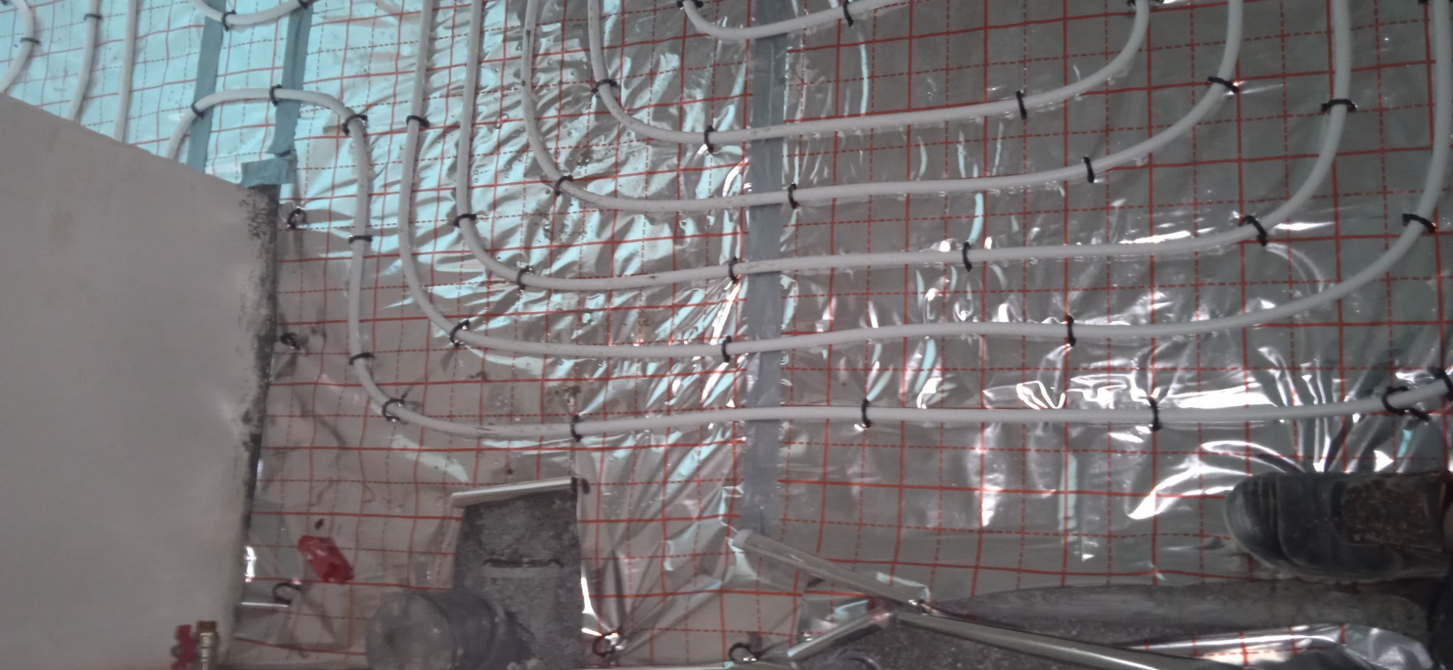 Białe rurki systemu ogrzewania podłogowego, mocowane czarnymi klipsami do folii izolacyjnej z czerwoną siatką, widok z góry na etapie instalacji.