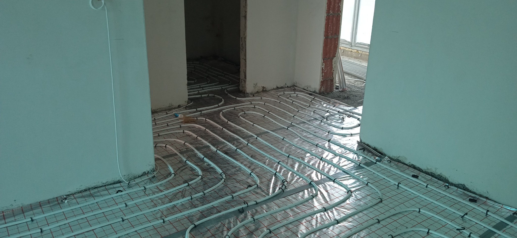 Białe rurki ogrzewania podłogowego ułożone na czerwonej siatce w surowym stanie, widoczne fragmenty ścian i przejścia do innych pomieszczeń.