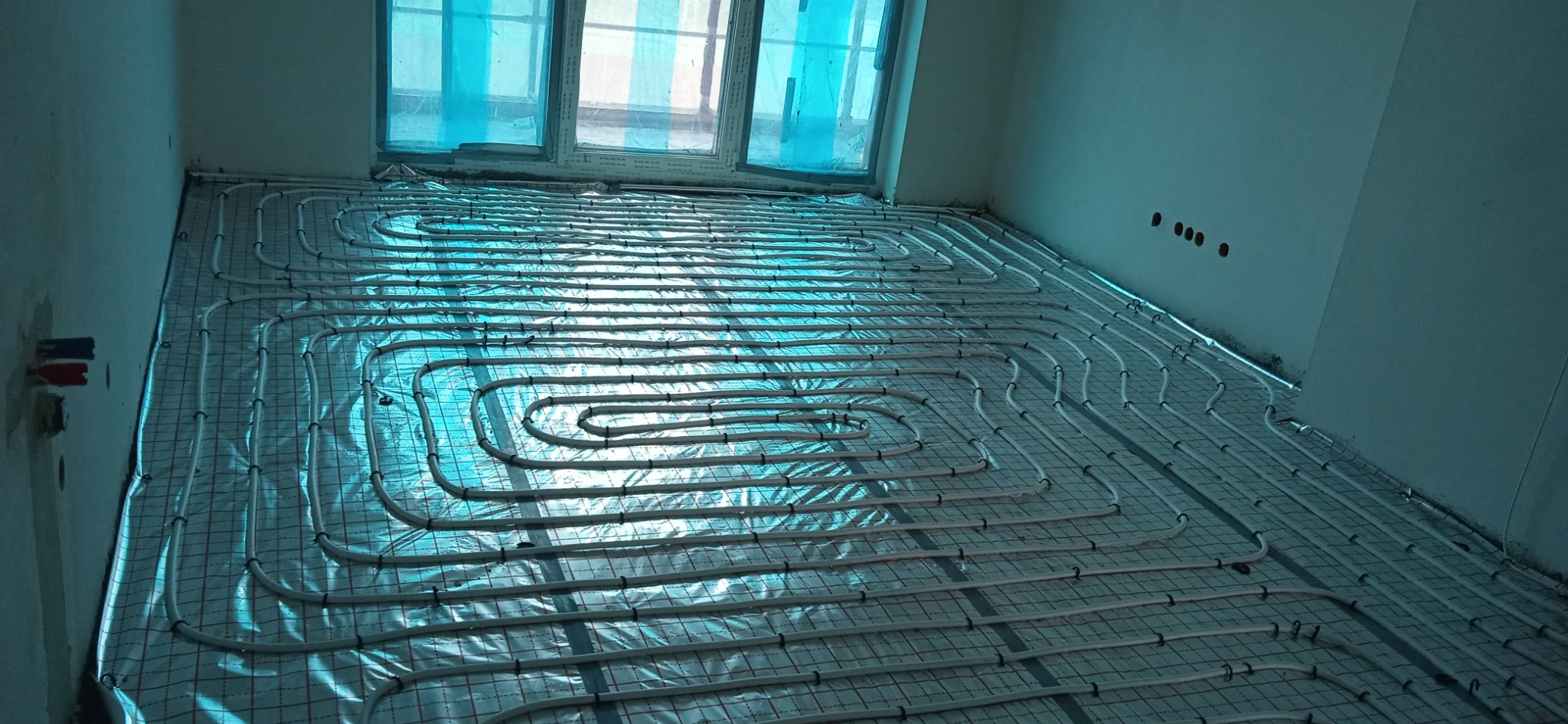 Białe rurki układane w pętle na siatce podłogowej, instalacja ogrzewania podłogowego w surowym pomieszczeniu z oknami zabezpieczonymi folią.