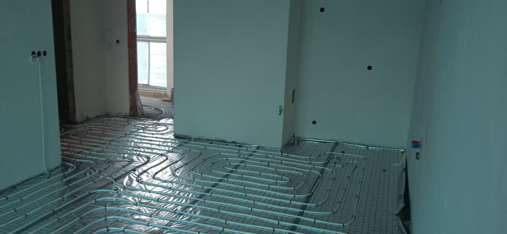 Rozłożone rury grzewcze na folii aluminiowej w trakcie montażu systemu ogrzewania podłogowego w surowym wnętrzu, widoczne wyprowadzenia elektryczne i ślady po wierceniu w ścianach.