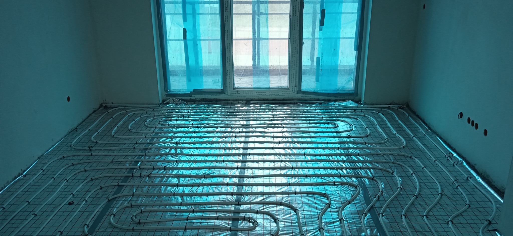 Białe rurki instalacji ogrzewania podłogowego ułożone na srebrnej folii izolacyjnej i metalowej siatce w surowym pomieszczeniu z oknem zabezpieczonym folią.