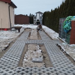 Vadym Smyrnyy - Układanie szarej ażurowej kostki brukowej na podjeździe w trakcie prac, widoczny pracownik klęczący przy układaniu i częściowo ułożona nawierzchnia, tło stanowi dom i ogród.