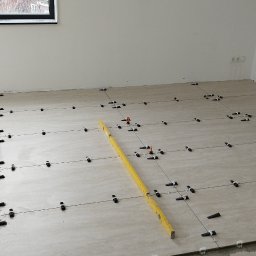 Vadym Smyrnyy - Układanie beżowych płytek podłogowych z użyciem systemu poziomowania, widoczna poziomica i krzyżyki dystansowe.