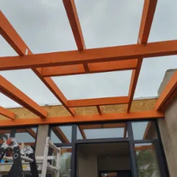 Pomarańczowa drewniana konstrukcja z belek i płyt OSB, widok od dołu na tle pochmurnego nieba i okien budynku, drabina oparta o ścianę.