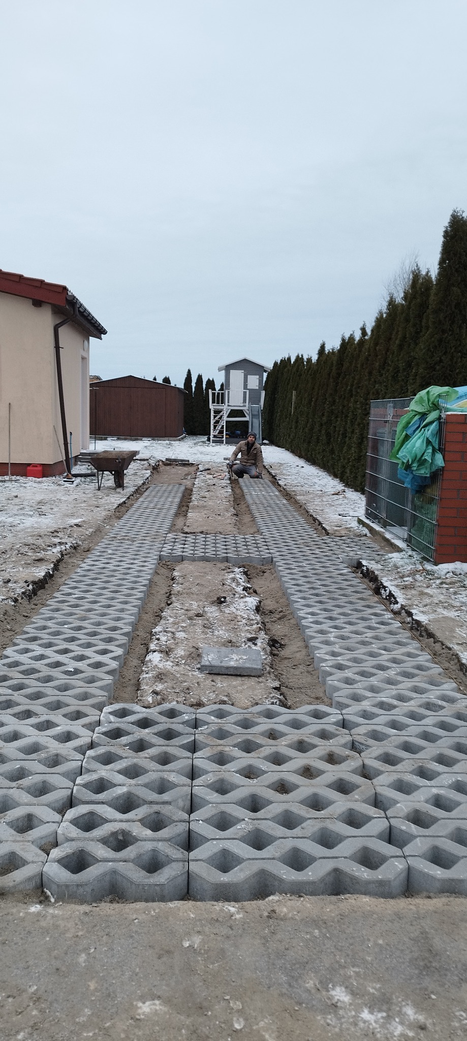 Układanie szarej ażurowej kostki brukowej na podjeździe w trakcie prac, widoczny pracownik klęczący przy układaniu i częściowo ułożona nawierzchnia, tło stanowi dom i ogród.