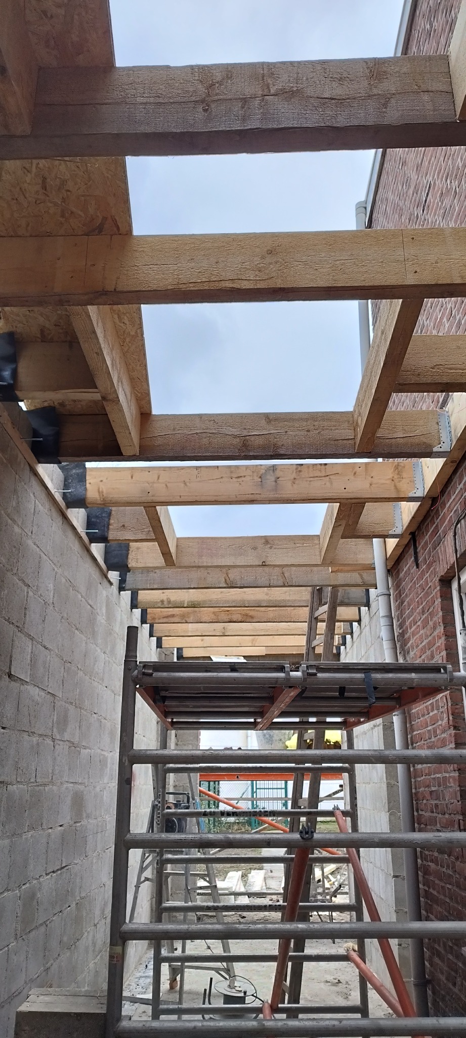 Konstrukcja drewniana stropu z widocznymi belkami i metalowymi łącznikami, w tle mur z cegły i rusztowanie.