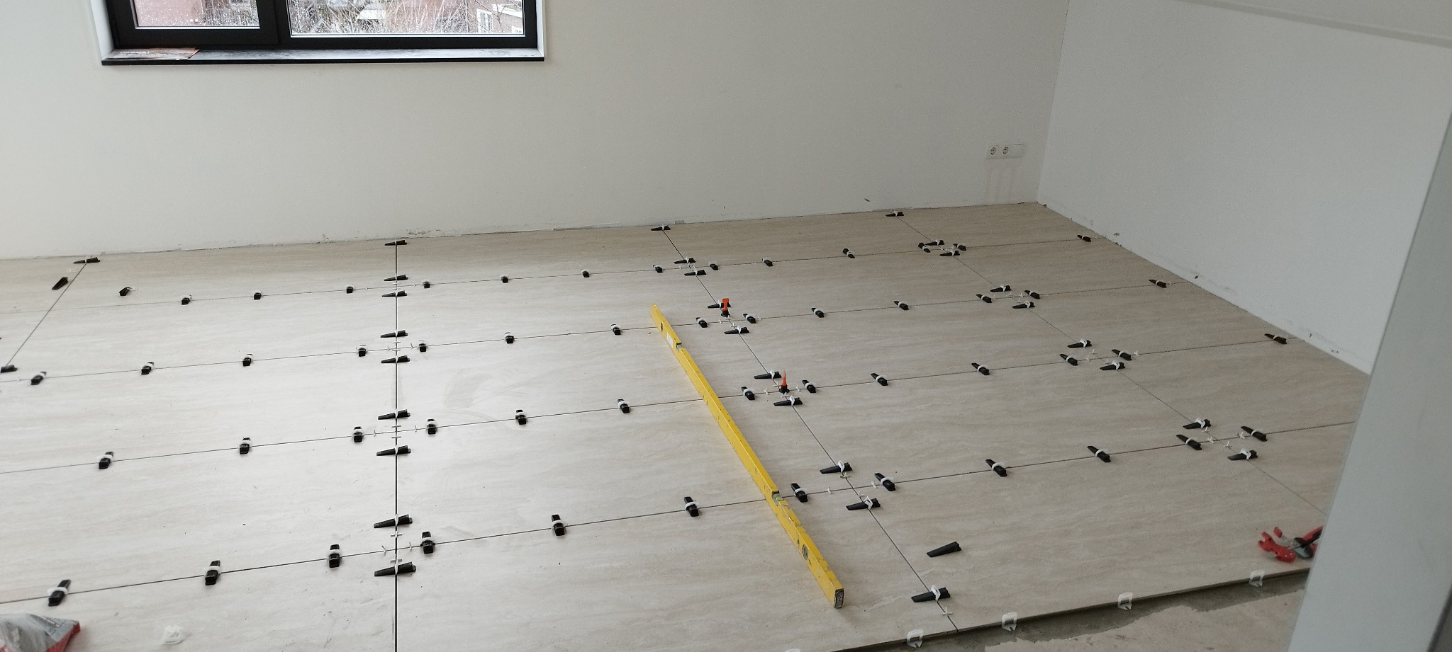 Układanie beżowych płytek podłogowych z użyciem systemu poziomowania, widoczna poziomica i krzyżyki dystansowe.