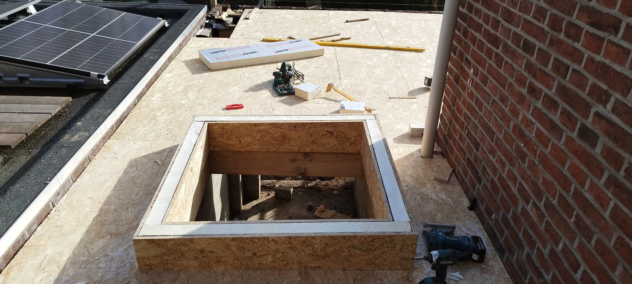 Montaż okna dachowego w trakcie prac: widoczny otwór w dachu z drewnianą konstrukcją, narzędzia pracy, płyty OSB, ceglana ściana i panel słoneczny w tle.