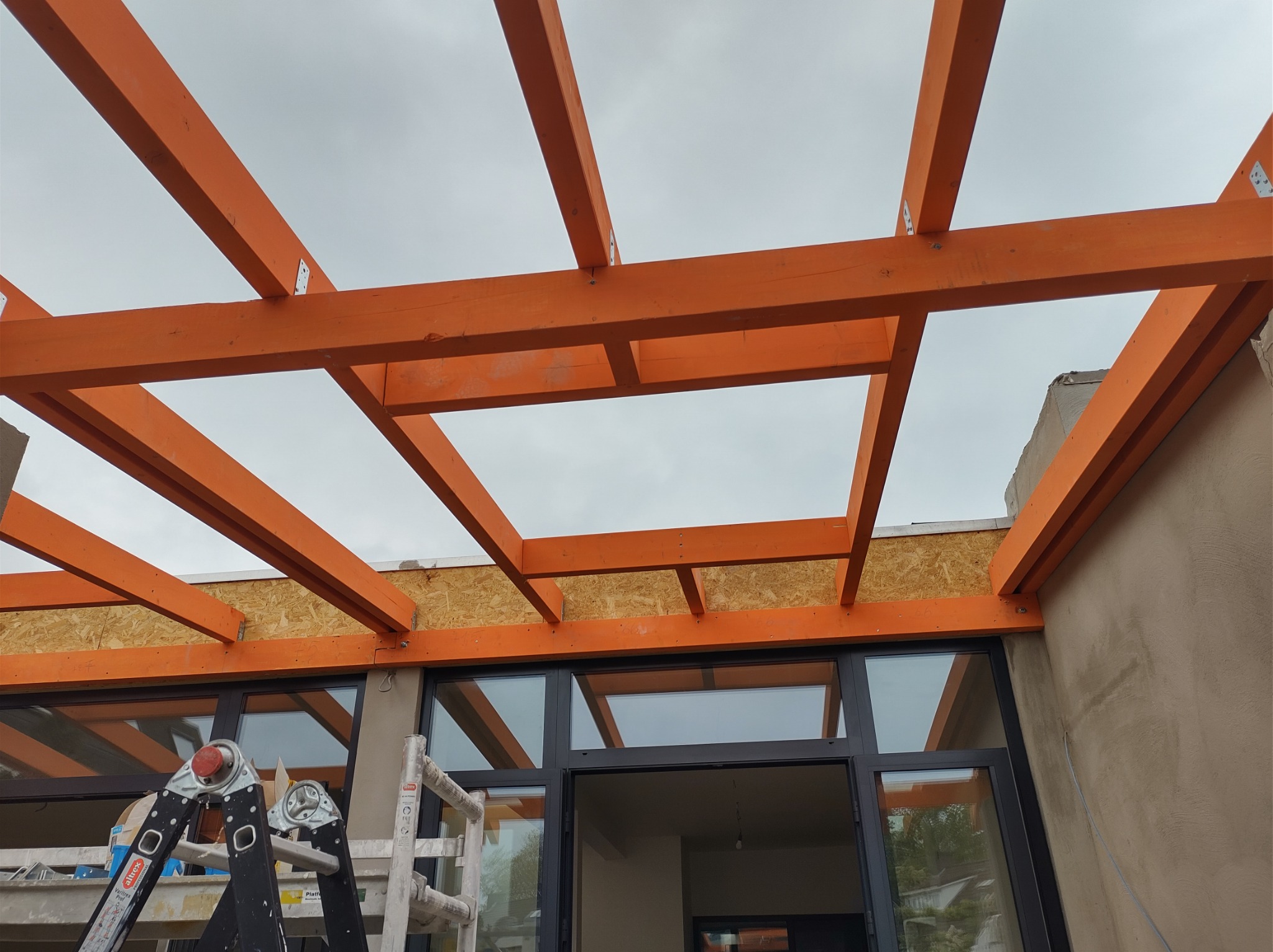 Pomarańczowa drewniana konstrukcja z belek i płyt OSB, widok od dołu na tle pochmurnego nieba i okien budynku, drabina oparta o ścianę.