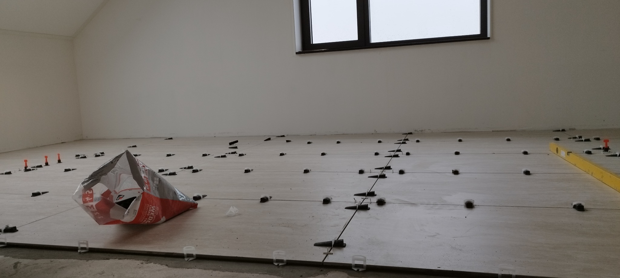 Układanie jasnych płytek podłogowych z użyciem systemu poziomowania, widoczny worek z klejem i poziomica.