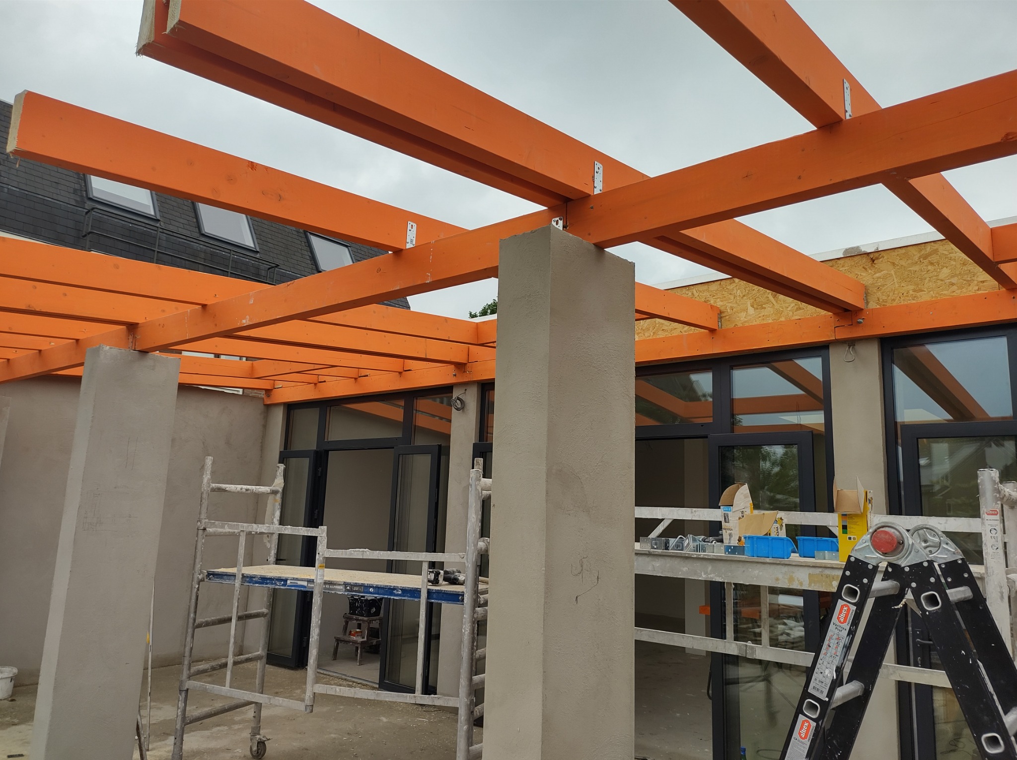 Montaż drewnianej konstrukcji z pomarańczowych belek na betonowych słupach, widoczne metalowe łączniki, rusztowania i okna w tle, budowa tarasu w trakcie realizacji.