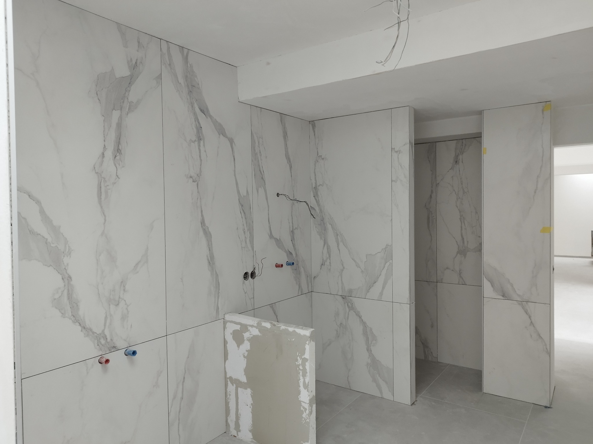 Wnętrze w trakcie wykańczania z marmurowymi płytkami na ścianach, widoczne wystające kable, fragment niedokończonej zabudowy i rury wodne.