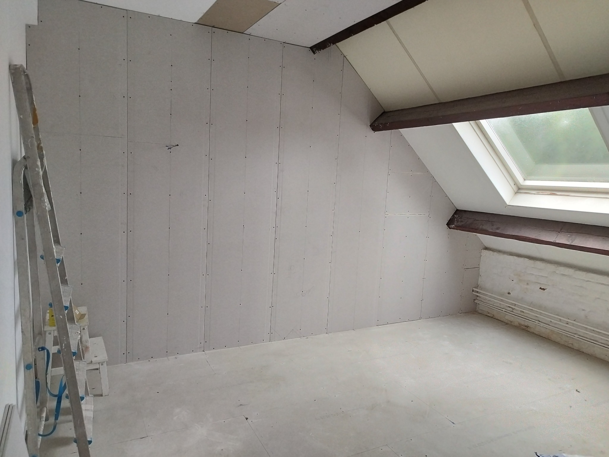 Montaż płyt karton gips na poddaszu ze skosami, widoczna drabina malarska i surowe drewniane belki stropowe.