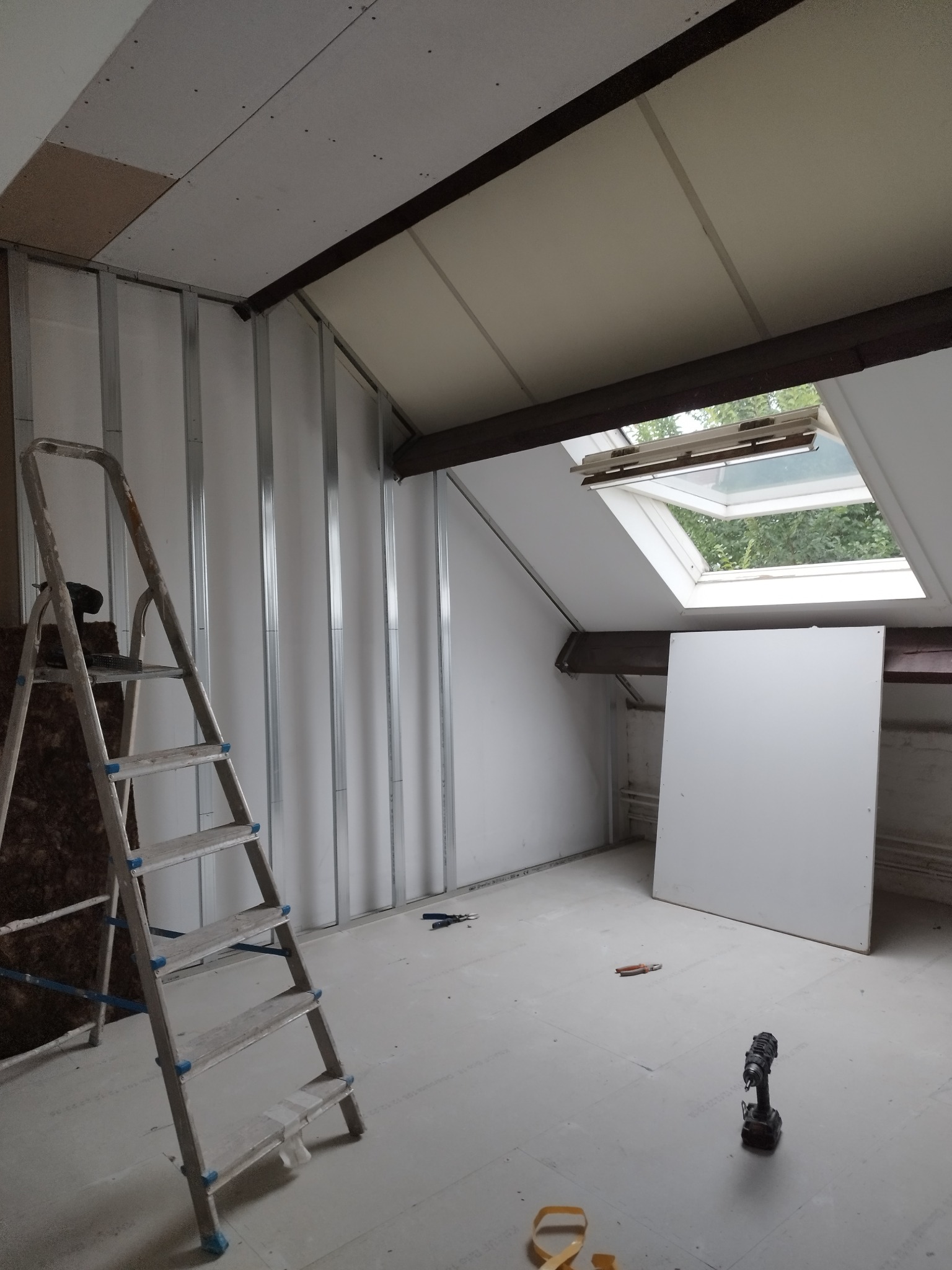 Poddasze w trakcie remontu: widoczne metalowe profile, częściowo zamontowane płyty gipsowo-kartonowe na ścianach, okno dachowe, drabina aluminiowa, wkrętarka na podłodze.