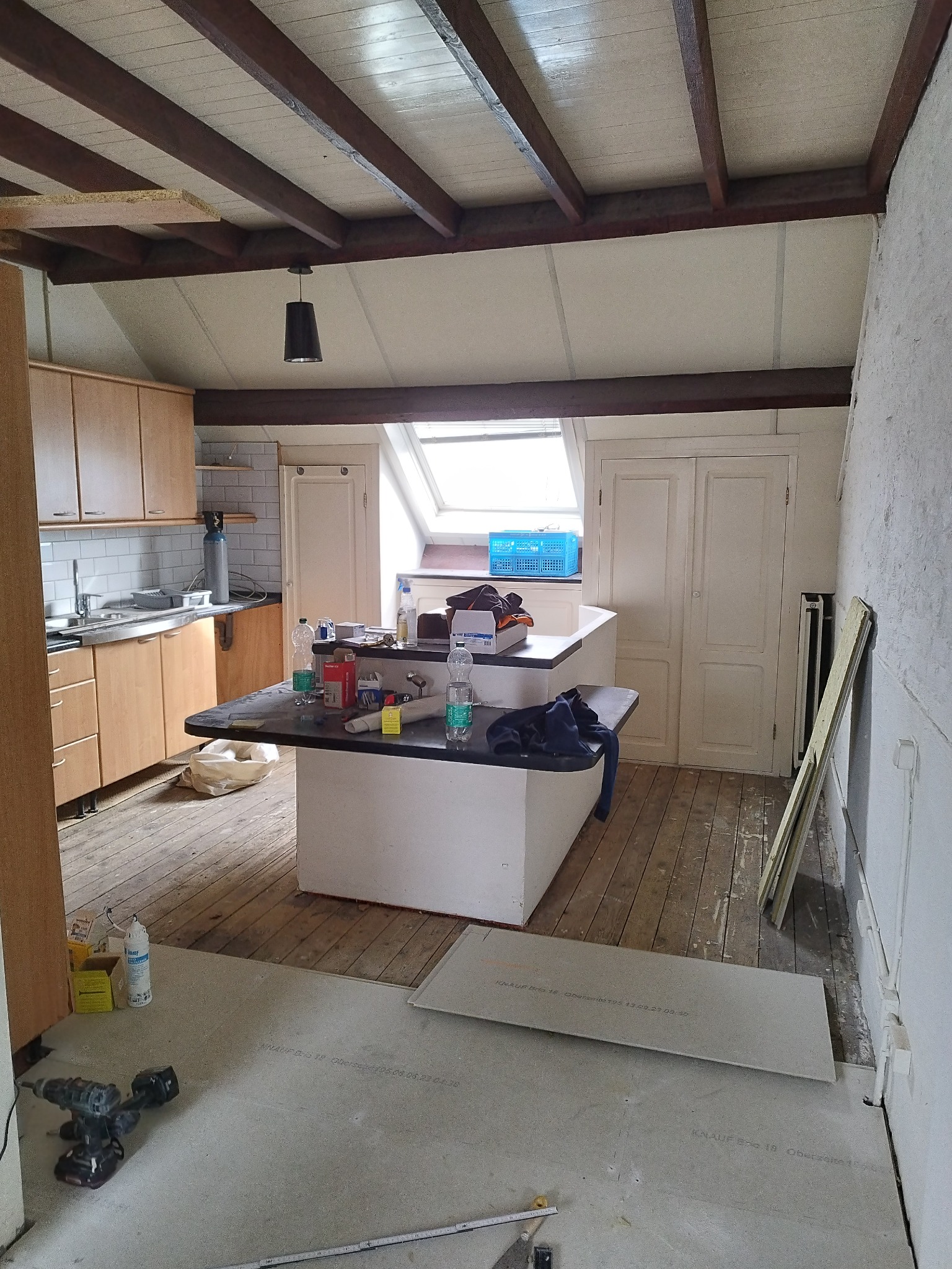 Wnętrze kuchni w trakcie remontu: widoczne płyty kartonowo-gipsowe na podłodze, drewniane belki sufitowe, zabudowa kuchenna i wyspa, narzędzia oraz materiały budowlane rozłożone na blacie.