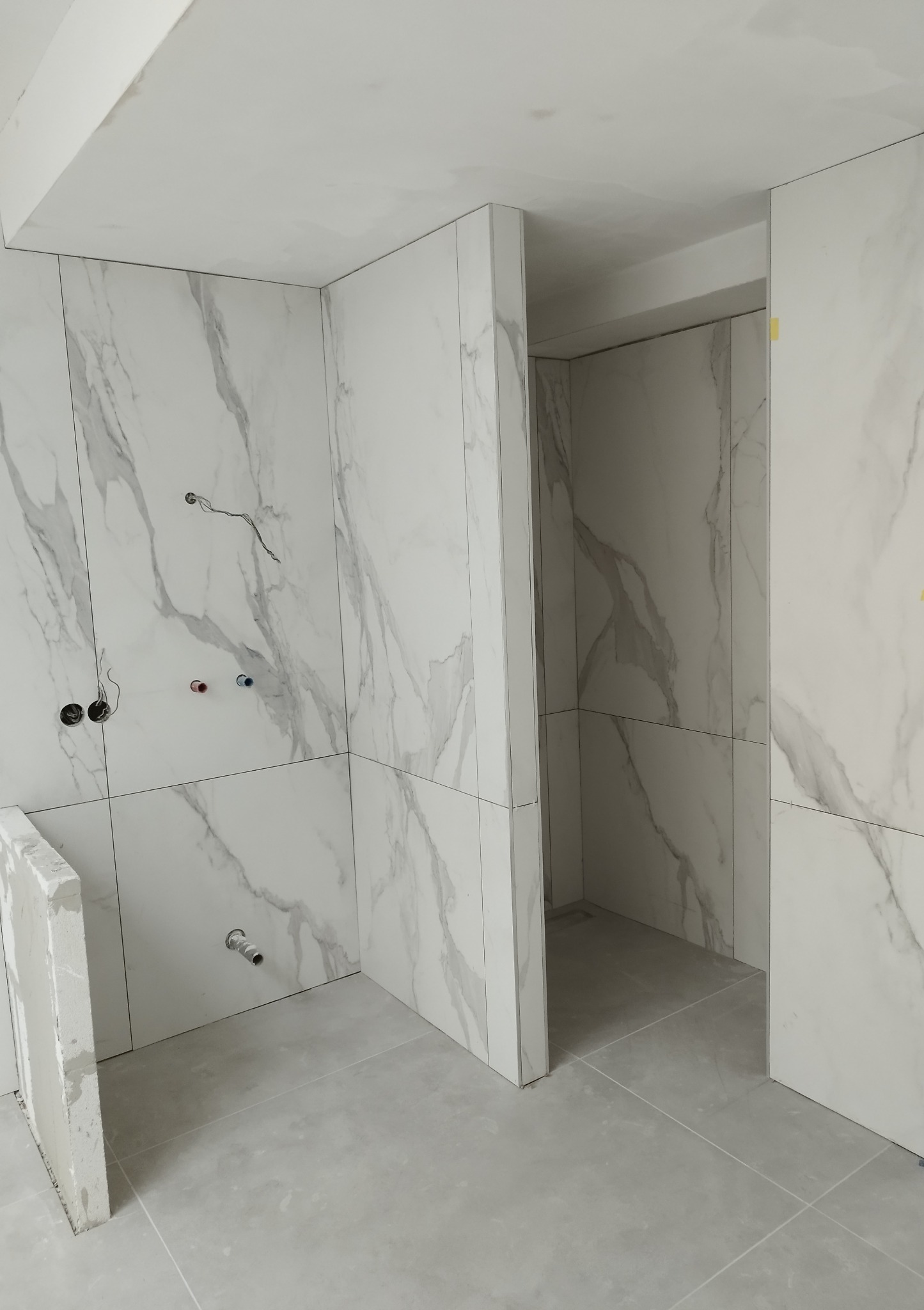 Wnętrze łazienki w trakcie wykańczania, widoczne płytki imitujące marmur na ścianach i podłodze, niezamaskowane przewody elektryczne i rury wodne, fragment ściany w stanie surowym.