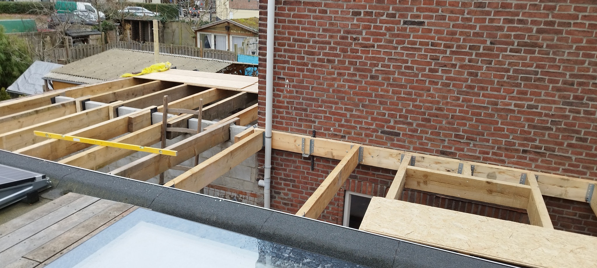 Konstrukcja drewniana dachu w trakcie budowy, widoczny poziomica, elementy mocujące i mur z czerwonej cegły.