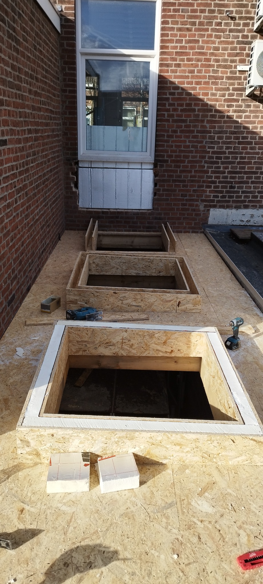 Przygotowanie otworów okiennych w dachu z OSB, obramowanych białą listwą, widoczne narzędzia i materiały izolacyjne.