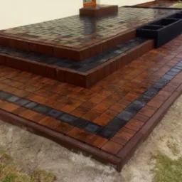 Ułożona kostka brukowa w odcieniach brązu i czerni tworząca schody i taras z donicą, widoczne ślady wilgoci po deszczu.