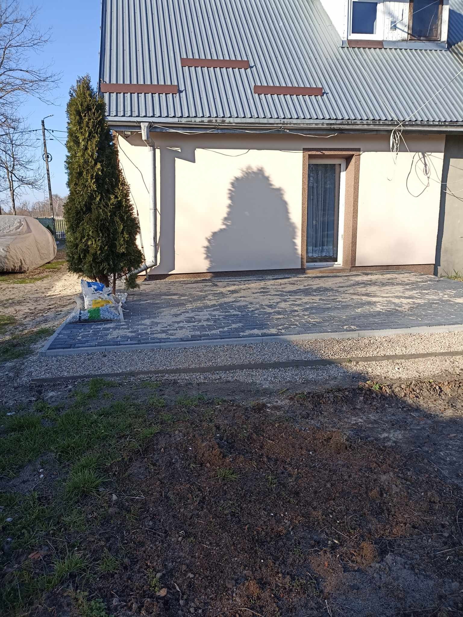 Ukończona nawierzchnia z szarej kostki brukowej przy jasnym domu z metalowym dachem, obok stoi worek z kamieniami i mała tuja.