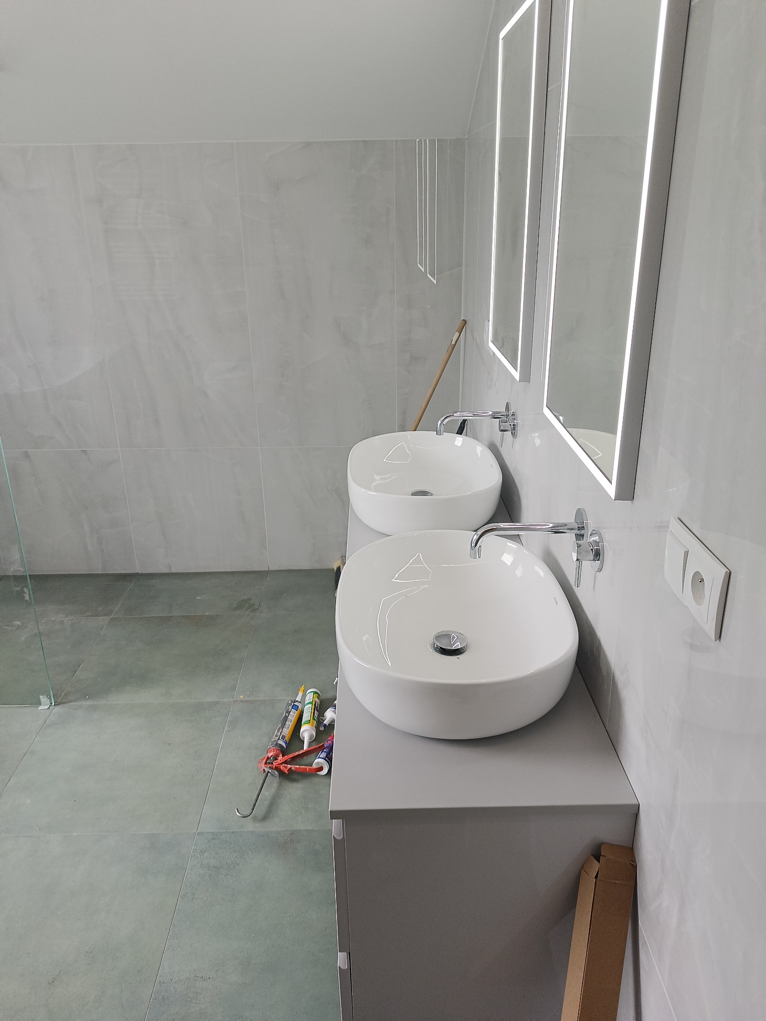 Dwie umywalki nablatowe na szarej szafce w łazience w Wesołej, podświetlane lustra, szare płytki ścienne i podłogowe, widoczne narzędzia i materiały budowlane.