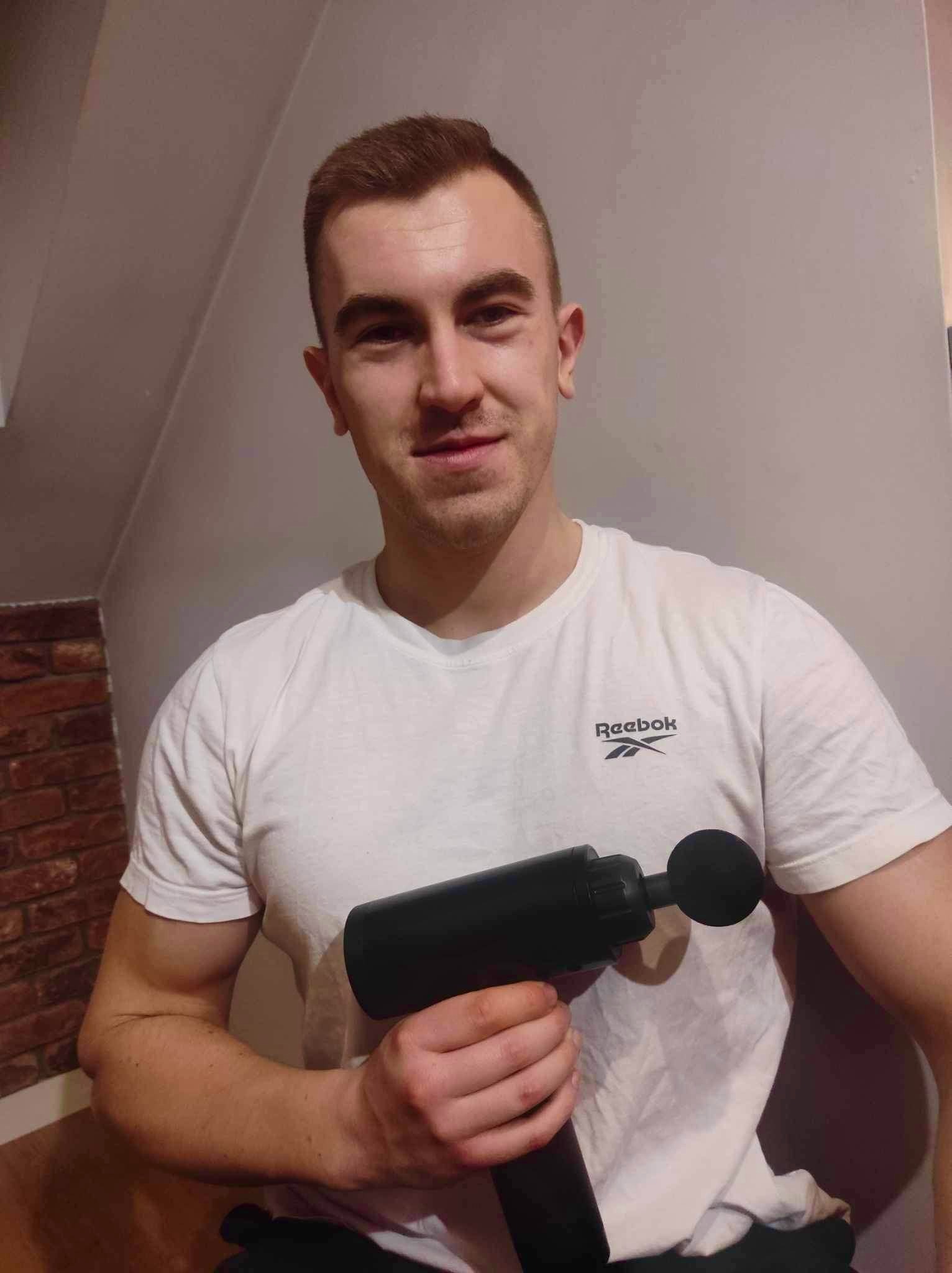 Mężczyzna w białej koszulce Reebok prezentuje czarny pistolet do masażu mięśni, stojąc na tle ściany z cegły i jasnej ściany.