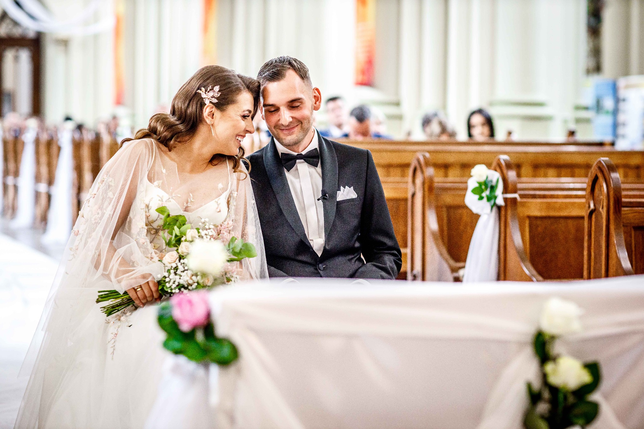 Uśmiechnięta panna młoda z bukietem i pan młody w czarnym smokingu siedzą blisko siebie w kościele, patrząc na siebie z czułością, w tle widoczne rzędy ławek z dekoracjami kwiatowymi.