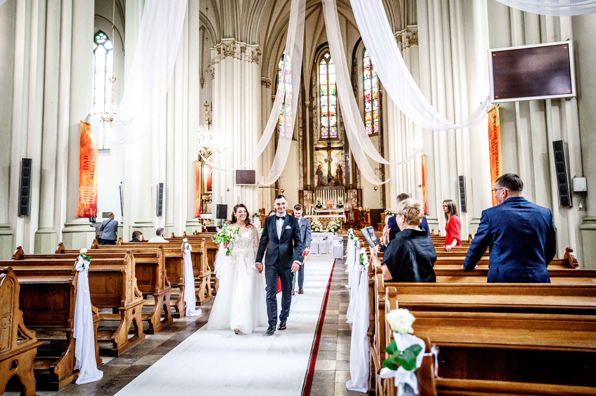 Para młoda, trzymając się za ręce, idzie wzdłuż białego dywanu w kościele, wśród gości siedzących w ławkach ozdobionych białymi kwiatami i wstążkami.