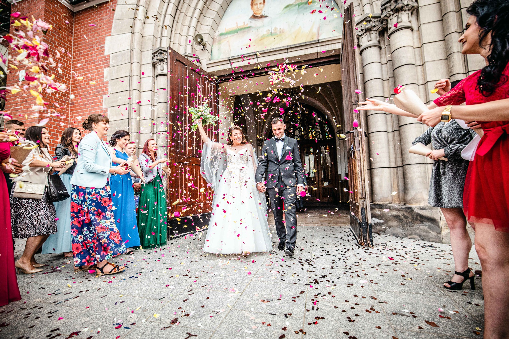 Para młoda opuszcza kościół w Zagrodach, obsypywana konfetti z płatków róż, panna młoda trzyma bukiet i unosi rękę w geście radości, na ścianie kościoła widoczny obraz religijny.