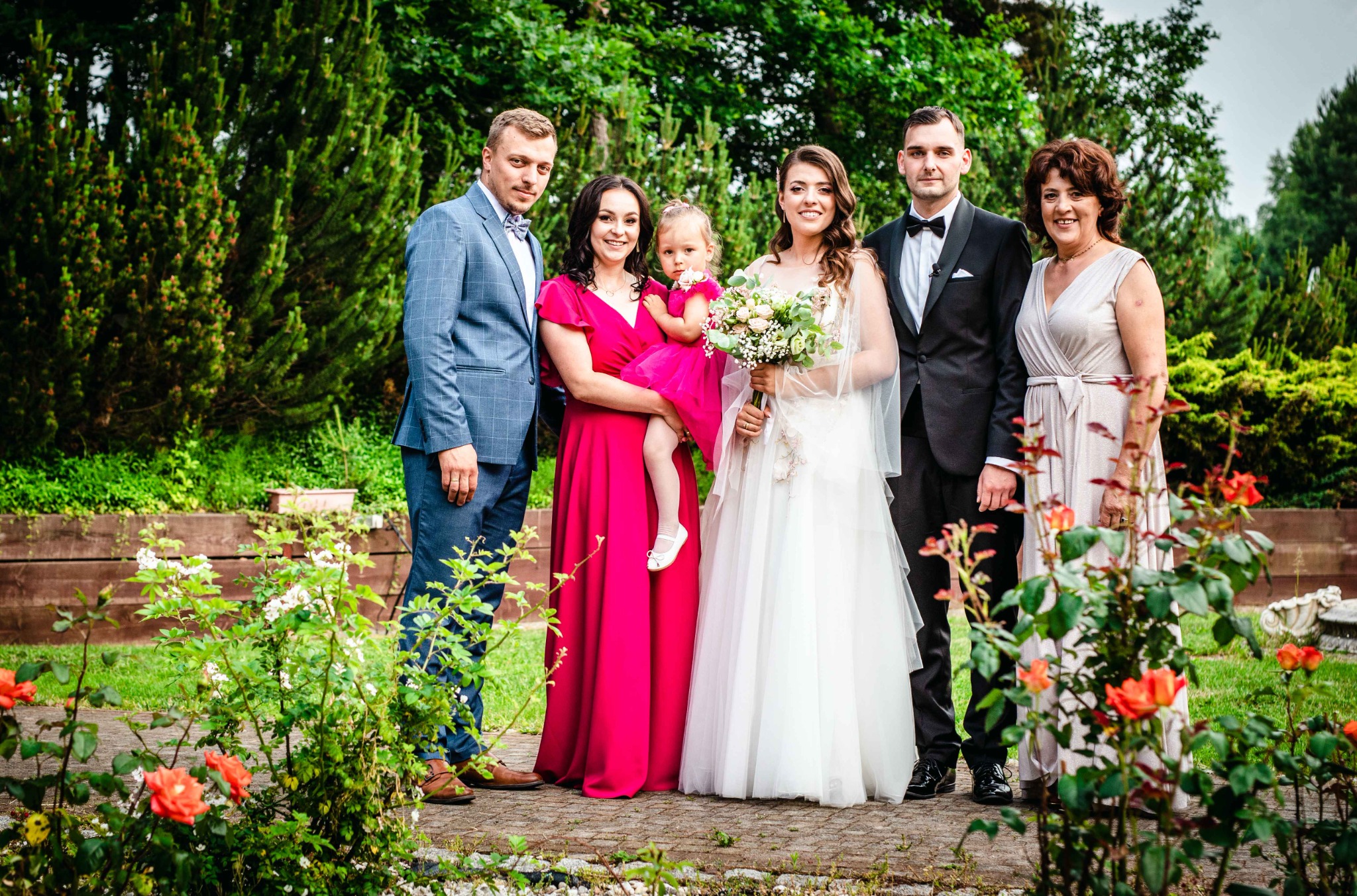 Grupa osób w strojach formalnych pozuje na zewnątrz, panna młoda trzyma bukiet kwiatów, w tle zielona roślinność i czerwone róże na pierwszym planie.