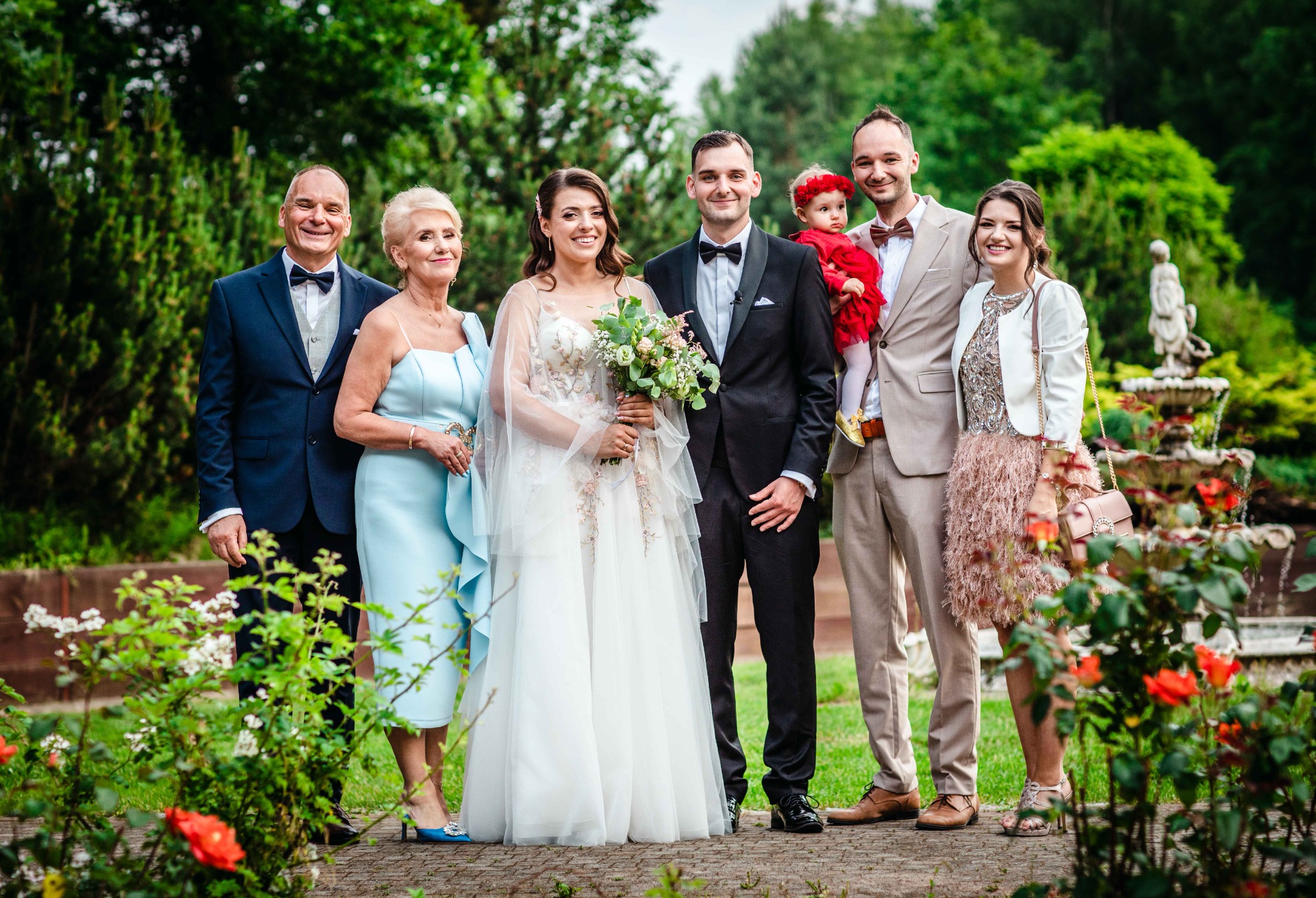 Uśmiechnięta para młoda z rodziną pozuje do zdjęcia w ogrodzie z fontanną. Panna młoda w białej sukni z haftowanymi kwiatami trzyma bukiet. Dziecko w czerwonej sukience na rękach jednego z mężczyzn.