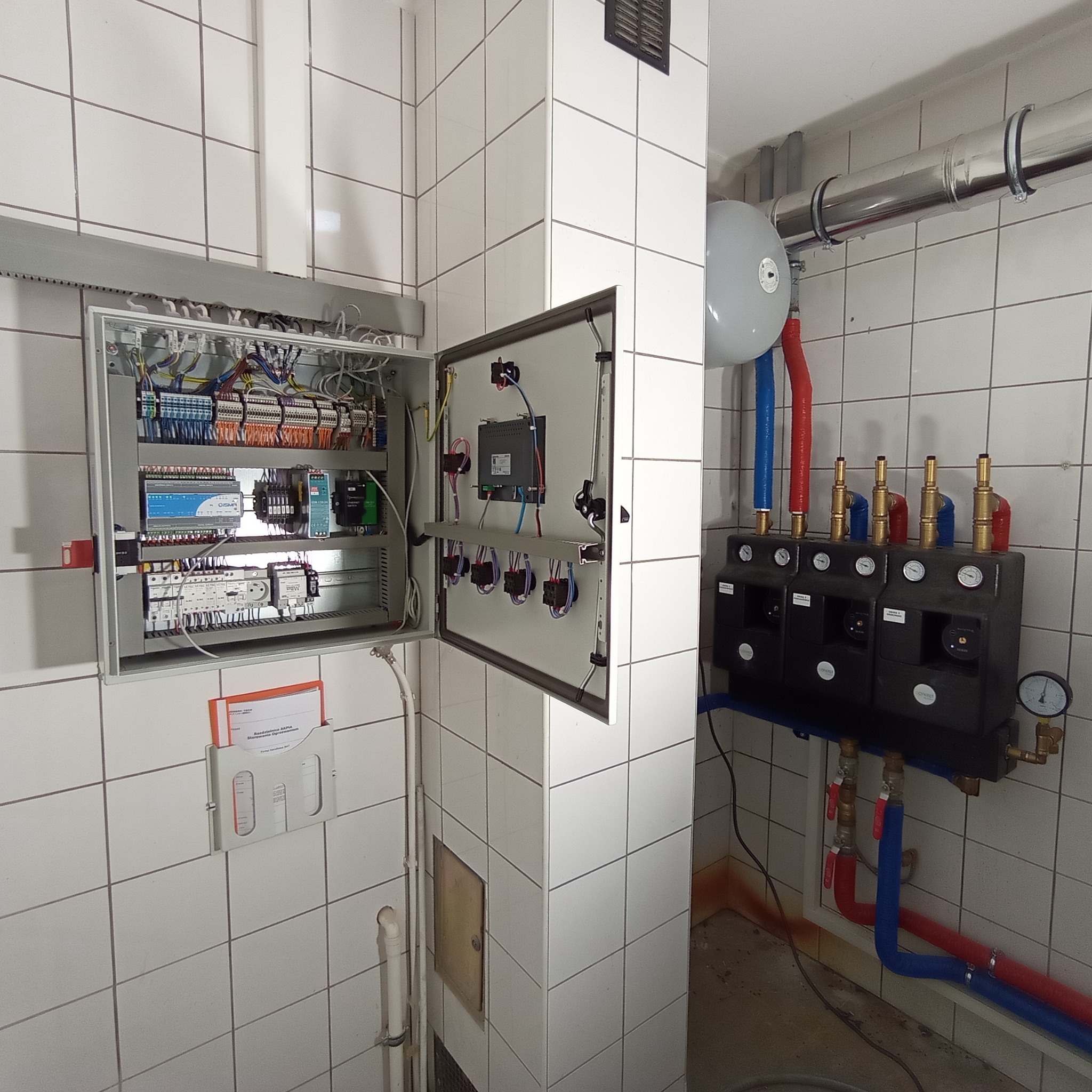 Otwarta szafa sterownicza z licznymi przewodami, bezpiecznikami i modułami, zamontowana na kafelkowanej ścianie obok kotłowni z rurami w izolacji czerwonej i niebieskiej oraz zbiornikiem wyrównawczym.